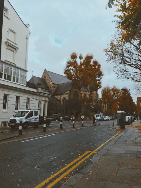 Photograph of a residential street in Stoke Newington during daytime, showing a row of parked cars along the curb with a white van belonging to Man with Van Stoke Newington parked adjacent to the sidewalk. The scene features a mix of white and darker vehicles, and the pavement is lined with black and white bollards. Tall trees with autumn foliage extend over the street, with some orange and brown leaves visible on the trees and scattered on the ground. In the background, there are historic buildings, including a stone church or similar structure with a steep roof and arched windows, partially obscured by the trees. The sky is overcast, contributing to a subdued, neutral lighting. This street scene illustrates a typical urban environment suitable for home relocation and furniture transport services, with the Man with Van Stoke Newington team potentially engaged in loading or unloading furniture during a moving process, emphasizing the logistics and outdoor environment involved in house removals in Stoke Newington.