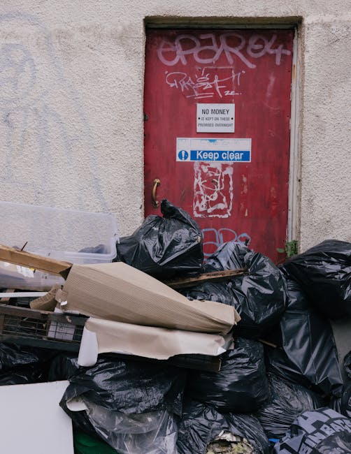 A red metal door with graffiti and signage, including a white 'No Money' notice and a blue 'Keep Clear' sticker, is set into a beige exterior wall. In front of the door, there is a pile of black rubbish bags filled with waste, some torn open revealing household packaging and discarded materials. Alongside the bags, there are flattened cardboard boxes, a plastic crate, and pieces of foam or packing material, possibly from packing during a home relocation or furniture transport. The scene appears to be at a loading area outside a property, with the rubbish awaiting disposal or collection. This image captures elements typical of moving or decluttering processes and highlights the importance of proper waste management during household moves, as managed by professional removal services like Man with Van Stoke Newington.