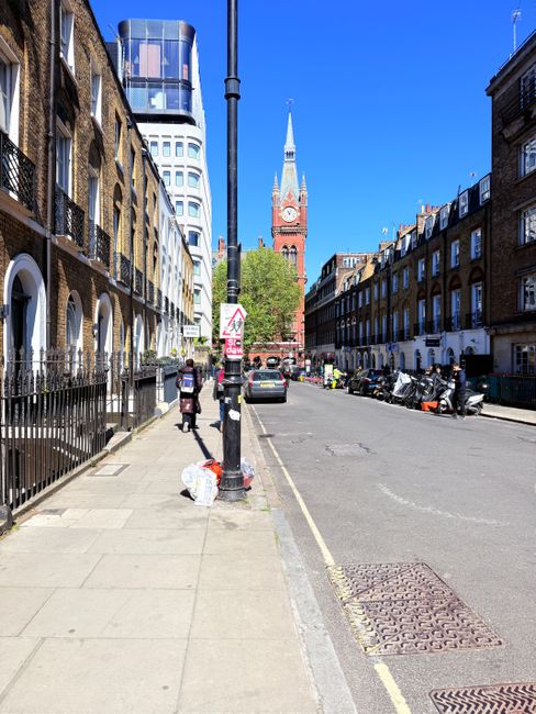 A wide city street scene in Stoke Newington with a clear blue sky, featuring residential buildings with brick facades and white window frames on both sides. On the left, there is a row of townhouses with black iron railings and small front gardens. A tall lamppost with a street sign is near the curb, where a person is seen walking while carrying a bag. Several cardboard boxes and plastic bags are placed on the sidewalk close to the lamppost, indicating preparatory packing for a home relocation. Parked motorcycles and a small car are visible along the side of the street, with a few pedestrians and vehicles in motion further ahead. In the background, the prominent spire of St. Mary's Church rises above the rooftops, and modern high-rise buildings with glass facades can be seen, contrasting with the older architecture. This scene illustrates the urban environment typical for local house removals and furniture transport services offered by Man with Van Stoke Newington during a moving or packing process.