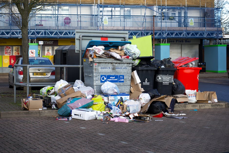 A red metal door with graffiti and signage, including a white 'No Money' notice and a blue 'Keep Clear' sticker, is set into a beige exterior wall. In front of the door, there is a pile of black rubbish bags filled with waste, some torn open revealing household packaging and discarded materials. Alongside the bags, there are flattened cardboard boxes, a plastic crate, and pieces of foam or packing material, possibly from packing during a home relocation or furniture transport. The scene appears to be at a loading area outside a property, with the rubbish awaiting disposal or collection. This image captures elements typical of moving or decluttering processes and highlights the importance of proper waste management during household moves, as managed by professional removal services like Man with Van Stoke Newington.