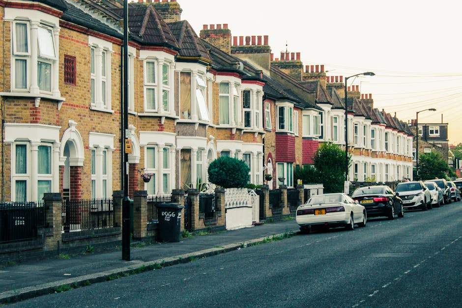A row of traditional terraced houses in Stoke Newington with brick facades and white bay windows, each with small front gardens enclosed by low brick and iron fences. The sidewalk runs along the front of the houses, with a black trash bin and a yellow blinking streetlight visible on the pavement. Several vehicles, including a white car, a black car, and a silver van, are parked parallel to the curb on the street. The image shows a typical residential street scene during daylight, with a clear sky and street lighting poles along the pavement. The scene illustrates the environment where home relocation or furniture transport might take place, and occasionally, [COMPANY_NAME] provides moving services in such areas.