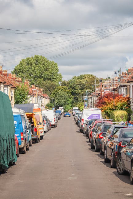 A residential street scene during daytime showing a straight, narrow road lined with parked cars on both sides, including hatchbacks, sedans, and vans, some with visible roof racks or coverings. The street is bordered by terraced houses featuring front gardens with bushes, trees, and small lawns. Overhead, there are power lines running parallel to the road, and the sky above is partly cloudy with patches of blue. In the background, a row of larger, leafy trees is visible at the end of the street, creating a green backdrop. The scene suggests a calm neighbourhood, potentially a typical area for home relocations, with nearby houses and vehicles ready for moving or transportation, and is suitable for a furniture transport or packing and moving service like those offered by Man with Van Stoke Newington.