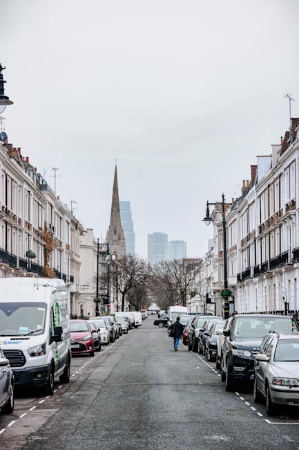 A narrow street in Stoke Newington lined with modern and older buildings on both sides, with some storefronts visible and a white van parked further down the road. The pavement is wet, suggesting rece
