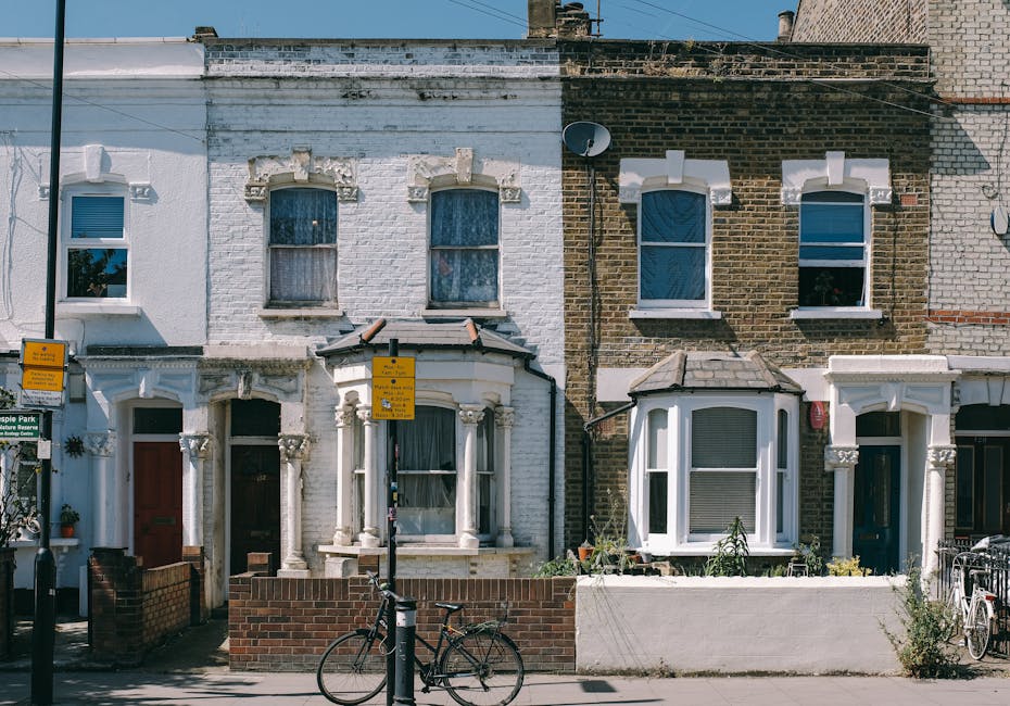A row of three terraced houses with brick and painted facades, each featuring a bay window on the ground floor and two smaller windows on the upper floors. The house on the left has a white exterior with decorative stonework around the door and window frames, and a small front garden enclosed by a low brick wall. The middle house has a white painted exterior with a bay window and a small roof canopy, with a black bicycle parked in front on the pavement. The house on the right has a brown brick facade with white decorative trim around the windows and a small front garden area with plants. The pavement in front is visible, with a yellow bus stop sign and a black lamppost in the foreground. The scene is lit with daylight, and the image relates to property move planning or home relocation services provided by Man with Van Stoke Newington, serving house removals and furniture transport needs in Stoke Newington.
