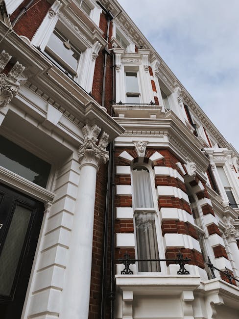 A low-angle view of a three-storey Victorian-style terraced house in Stoke Newington, featuring decorative white stone moldings, ornate columns with Corinthian capitals supporting the upper floors, and arched windows with white frames and sills. The building's façade combines red brickwork with contrasting white detailing, including horizontal bands and window surrounds. A small balcony with black wrought iron railings extends from the second-floor window, while the ground floor has a large glass entrance door framed by white brickwork. The sky overhead is cloudy, and the surrounding area suggests an urban residential setting. This image is relevant to house removals and furniture transport services, with [COMPANY_NAME] often assisting with packing, loading, and moving processes within such ornate properties.