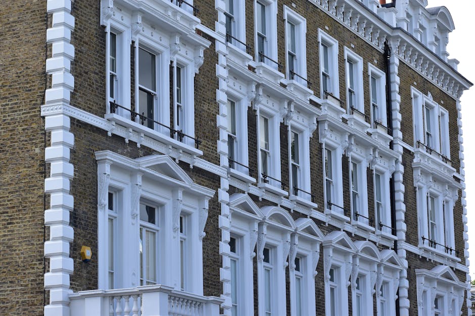 A row of three terraced houses with brick and painted facades, each featuring a bay window on the ground floor and two smaller windows on the upper floors. The house on the left has a white exterior with decorative stonework around the door and window frames, and a small front garden enclosed by a low brick wall. The middle house has a white painted exterior with a bay window and a small roof canopy, with a black bicycle parked in front on the pavement. The house on the right has a brown brick facade with white decorative trim around the windows and a small front garden area with plants. The pavement in front is visible, with a yellow bus stop sign and a black lamppost in the foreground. The scene is lit with daylight, and the image relates to property move planning or home relocation services provided by Man with Van Stoke Newington, serving house removals and furniture transport needs in Stoke Newington.