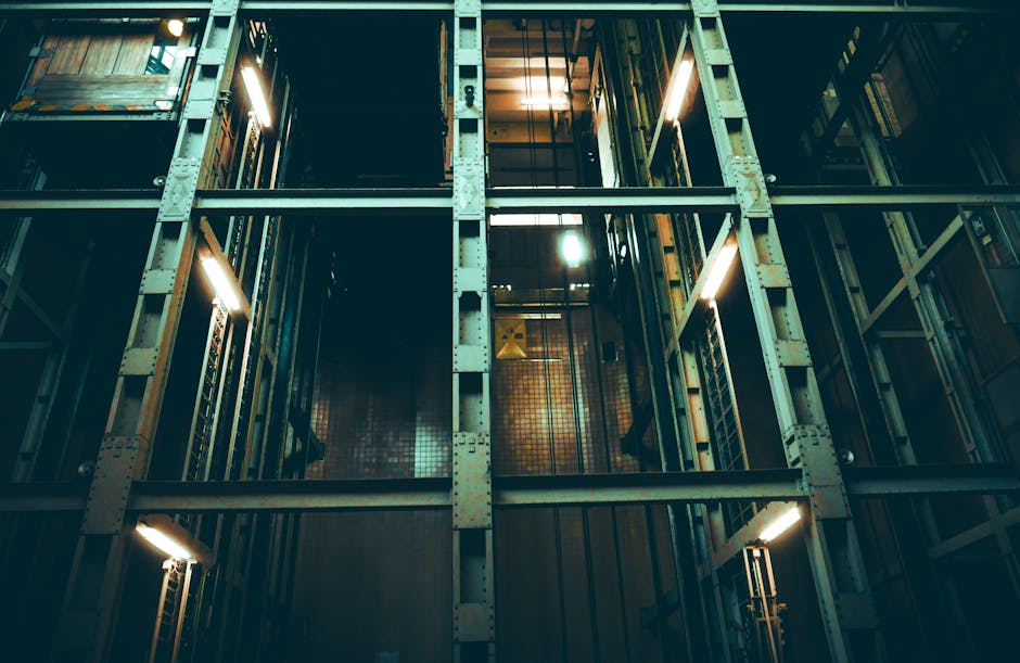 The image depicts a view of the interior of a multi-storey building's stairwell or lift shaft, showing metal scaffolding and beams illuminated by fluorescent lighting. The setting appears industrial, with visible support structures and open sections revealing the dark space beneath. The area is inside a building, likely involved in a house removal or furniture transport process, with the structure prepared for moving equipment or lighting to facilitate access for packing and moving activities. There are no visible objects or items being moved directly in the image, but the environment suggests ongoing preparations or logistics typical of professional removals by companies like Man with Van Stoke Newington. The lighting emphasizes the vertical and horizontal supports, highlighting the structural framework used during home relocation projects, especially in buildings with narrow access points or stairwells requiring careful navigation during furniture transport.