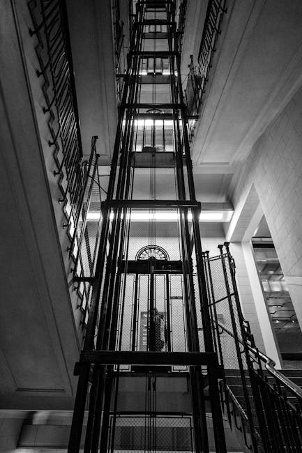 Interior view of a building's central stairwell with a modern, black metal lift platform visibly installed within the structure. The lift extends vertically from the ground floor to upper levels, with metal rails and steps clearly shown, designed to facilitate the transport of furniture and belongings during home relocation. The surrounding walls are tiled or paneled in light-colored materials, with a large window at the top providing natural light. The lift is positioned adjacent to a staircase, which is partially visible on the side, and the area appears to be part of a residential or commercial property preparing for a moving process. This setting illustrates the use of specialized equipment by Man with Van Stoke Newington for navigating narrow lifts during furniture transport and packing and moving operations.