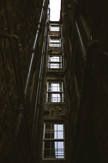 The image depicts a view of the interior of a multi-storey building's stairwell or lift shaft, showing metal scaffolding and beams illuminated by fluorescent lighting. The setting appears industrial, with visible support structures and open sections revealing the dark space beneath. The area is inside a building, likely involved in a house removal or furniture transport process, with the structure prepared for moving equipment or lighting to facilitate access for packing and moving activities. There are no visible objects or items being moved directly in the image, but the environment suggests ongoing preparations or logistics typical of professional removals by companies like Man with Van Stoke Newington. The lighting emphasizes the vertical and horizontal supports, highlighting the structural framework used during home relocation projects, especially in buildings with narrow access points or stairwells requiring careful navigation during furniture transport.