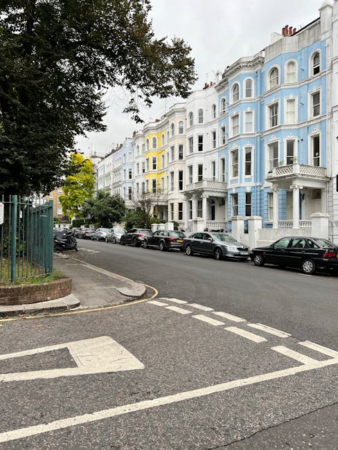 A view of a residential street in Stoke Newington featuring a row of pastel-coloured Victorian-style terraced houses with bay windows and decorative facades, painted mainly in light blue, white, and yellow. Several parked cars line the curb, including a mix of compact and larger vehicles, on the right side of the street. The road surface is asphalt, with a pedestrian crossing marked by white lines and a small traffic island or curb in the foreground. To the left, a large leafy tree with dark green foliage partially obscures the view of the houses and casts shadows over part of the street. A green metal fence borders a landscaped area with some plants and a brick base. The sky is overcast, creating diffuse lighting conditions. This scene depicts an urban environment suitable for residential house removals and furniture transport, as handled by companies like Man with Van Stoke Newington, who manage packing, loading, and parking logistics in busy neighbourhoods.