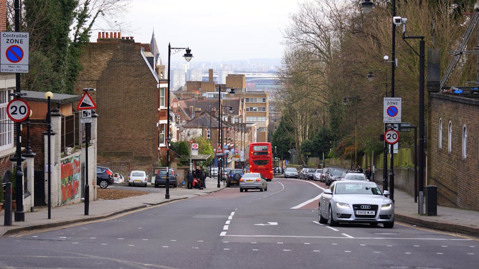 A street scene in Stoke Newington featuring parked vehicles along the sides of a curving road with a red double-decker bus in the distance. On the left side, there are several cars parked alongside the pavement, with a group of people visible near a bus stop. Street signs indicating parking restrictions and speed limits are mounted on poles, including a controlled zone sign and a no parking zone. The buildings lining the street are a mix of residential houses and brick structures, some with small front gardens and others with external stairs. Street lamps and trees are present along the sidewalks, and in the background, there is a view of a city skyline with taller buildings under a partly cloudy sky. This scene illustrates typical urban traffic and parking arrangements, relevant for furniture transport, home relocation, and moving services provided by Man with Van Stoke Newington, especially regarding parking regulations and potential fine risks for N16 removals.