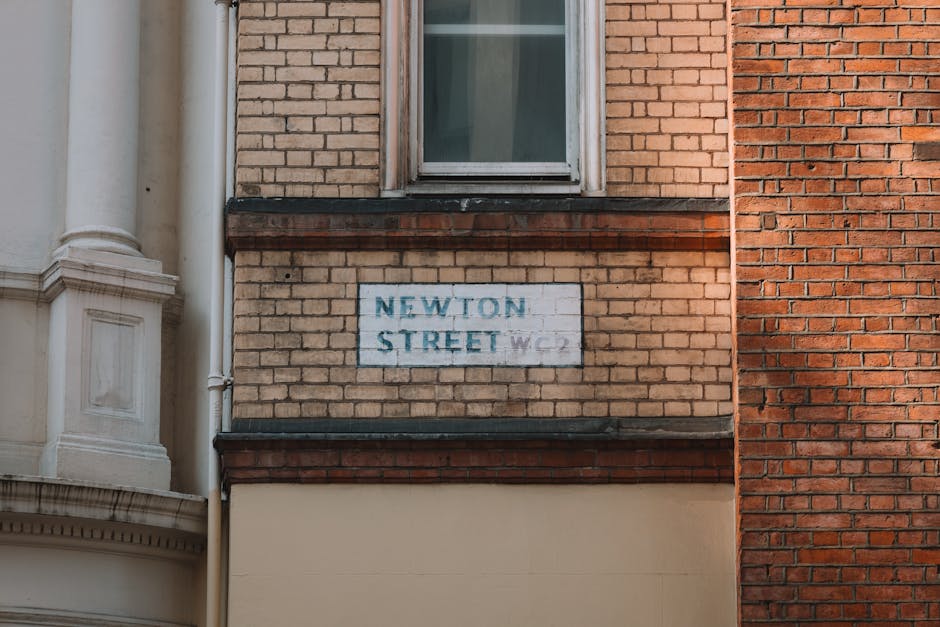 A close-up view of a brick building facade on Newton Street, displaying a small rectangular street sign with faded blue text that reads 'NEWTON STREET' and a smaller line beneath with additional characters, partially obscured. The sign is mounted on the lower part of the building's exterior wall, which features a combination of light beige, cream, and reddish-brown bricks. Above the sign, there is a closed window with a white frame. To the left, part of an ornate white column and decorative molding are visible, indicating the neighboring building. The image likely depicts a residential area in Stoke Newington, suitable for house removal or furniture transport projects. The setting suggests an urban environment where packing, loading, and moving logistics are involved, with the sign serving as a local reference point for relocation services provided by Man with Van Stoke Newington. The lighting appears natural, highlighting the textures of the bricks and the structural elements, emphasizing the building's architectural details relevant for home relocation planning.