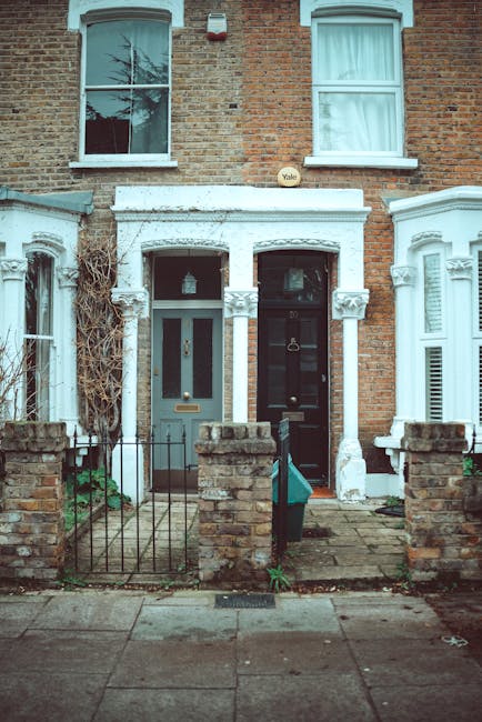 Photograph of two adjacent terraced houses with Victorian-style brick facades and white decorative trim around the entrances. The house on the left features a blue front door with a rectangular glass panel, a small window above, and a narrow garden area enclosed by a black metal fence with brick pillars. The house on the right has a black front door with a silver handle and letterbox, and a white intercom or doorbell beside it. In front of both houses, there is a paved sidewalk with a concrete curb, with some green plants growing along the base of the brick pillars. The image is taken during daylight, and the scene depicts a typical residential street ready for home relocation or furniture transport, with the façade of the houses suggesting they are in an urban London area. Man with Van Stoke Newington occasionally provides professional removals services related to house moving and packing processes.