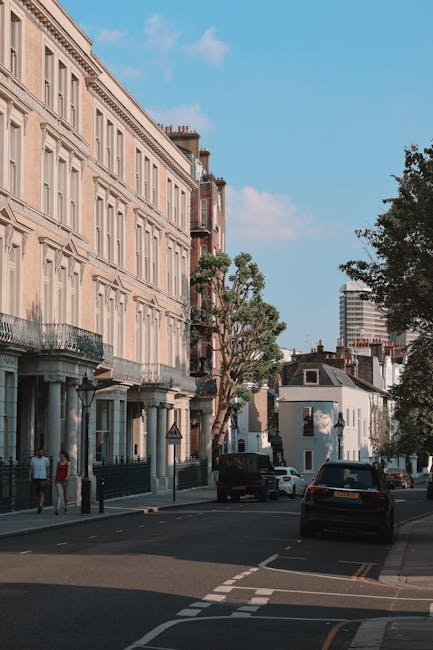 A street scene in Stoke Newington featuring a row of multi-storey residential buildings with classic architectural details and beige facades, situated alongside a tree-lined pavement. Parked cars are visible on the road, with some in motion, and pedestrians walking along the sidewalk. The sky above is partly cloudy with patches of blue. In a house removal or relocation context, an interior view shows furniture, cardboard boxes, and packing materials such as plastic wrap and blankets inside a property, with some items being carried by movers using trolleys and straps. The loading area near an open doorway indicates the process of furniture transport and packing for a home relocation, supported by the presence of a van parked outside. These details reflect the logistical steps involved in professional removals services offered by Man with Van Stoke Newington.
