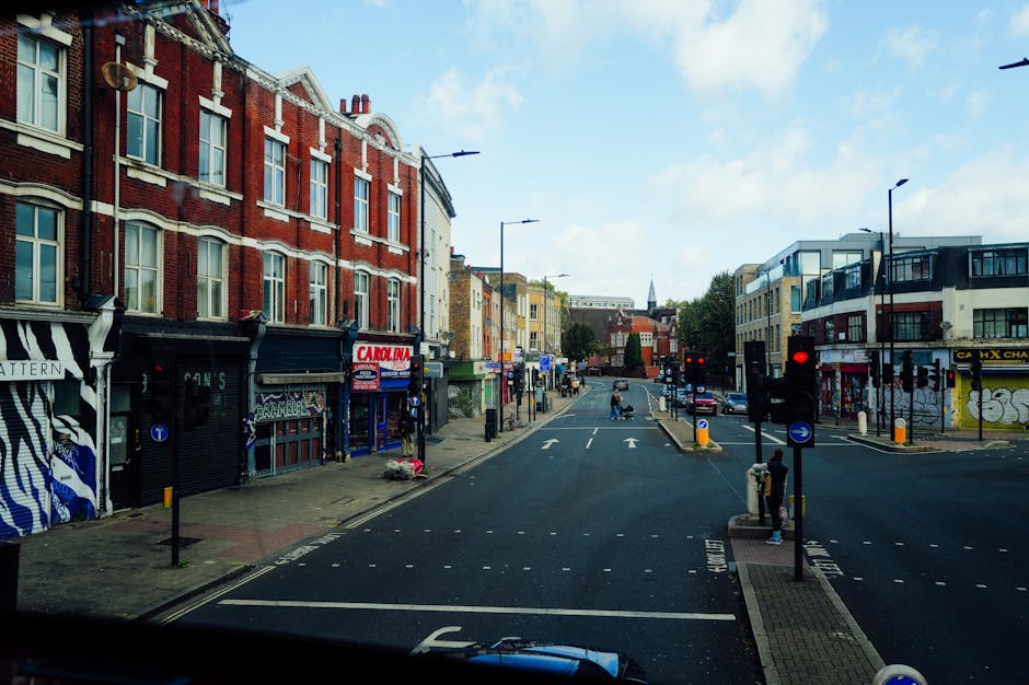 A street scene in Stoke Newington featuring a row of multi-storey residential buildings with classic architectural details and beige facades, situated alongside a tree-lined pavement. Parked cars are visible on the road, with some in motion, and pedestrians walking along the sidewalk. The sky above is partly cloudy with patches of blue. In a house removal or relocation context, an interior view shows furniture, cardboard boxes, and packing materials such as plastic wrap and blankets inside a property, with some items being carried by movers using trolleys and straps. The loading area near an open doorway indicates the process of furniture transport and packing for a home relocation, supported by the presence of a van parked outside. These details reflect the logistical steps involved in professional removals services offered by Man with Van Stoke Newington.